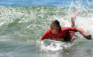 A man in a red rash guard lies on a surfboard, paddling through a breaking wave with splashing white water.